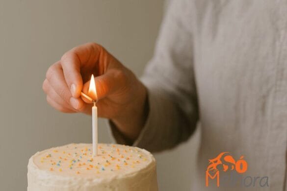 Mão acendendo uma vela de aniversário em um bolo branco decorado com confeitos coloridos.